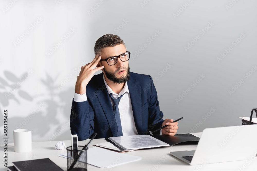 Tired businessman holding pen near documents, papers and laptop on blurred foreground in office Tired businessman holding pen near documents, papers and laptop on blurred foreground in office
