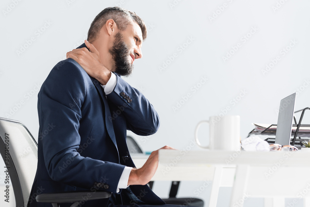 Businessman in formal wear suffering from neck pain near laptop and cup on blurred foreground in office Businessman in formal wear suffering from neck pain near laptop and cup on blurred foreground in office