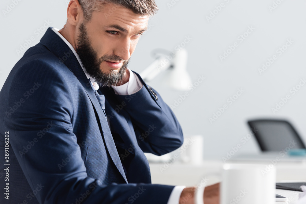 Obraz premium Businessman suffering from neck pain near cup on blurred foreground in office