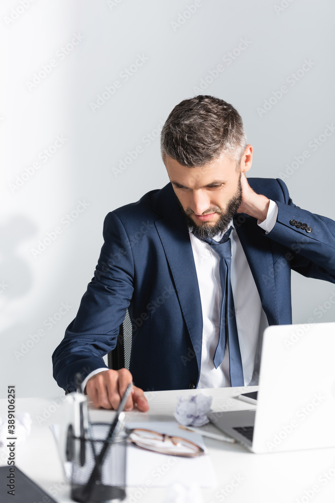 Businessman touching neck near laptop and stationery on blurred foreground in office