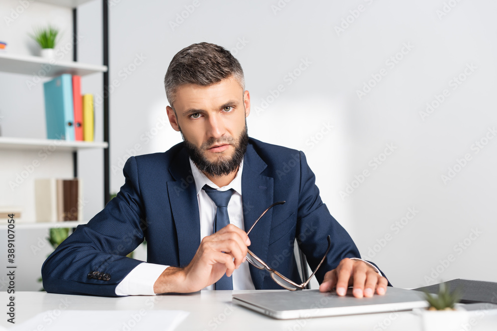 Businessman holding eyeglasses and looking at camera near laptop on blurred foreground in office Businessman holding eyeglasses and looking at camera near laptop on blurred foreground in office