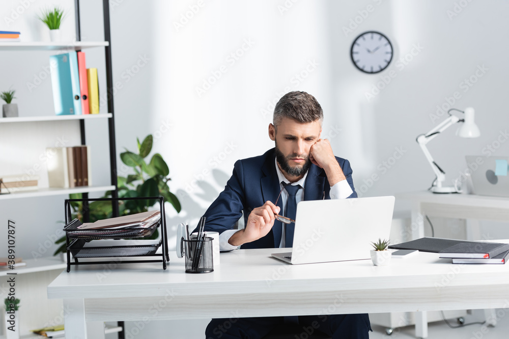 Businessman holding eyeglasses while looking at laptop near papers on table