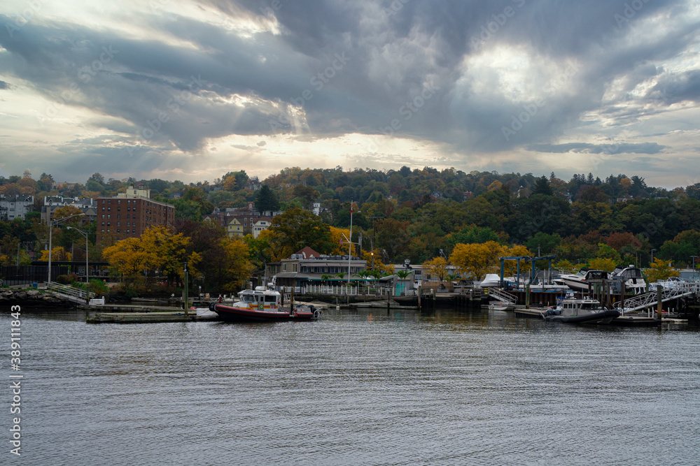 A view of a vast lake or river with several boats parked next to its coast with a dense forest visible in the distance under the cloudy autumn sky
