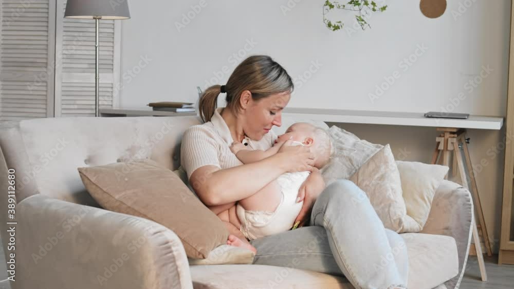 Medium shot portrait of young Caucasian woman sitting on sofa, watching toddler sleeping in her hands, then kissing him on nose and looking at camera Medium shot portrait of young Caucasian woman sitting on sofa, watching toddler sleeping in her hands, then kissing him on nose and looking at camera