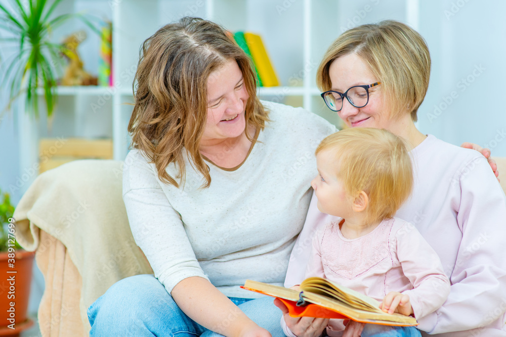 Two young women are sitting in the living room on the couch with a child on their knees and are reading a book to the child. Gay lesbian couple at home with a baby. Lesbian family concept Two young women are sitting in the living room on the couch with a child on their knees and are reading a book to the child. Gay lesbian couple at home with a baby. Lesbian family concept