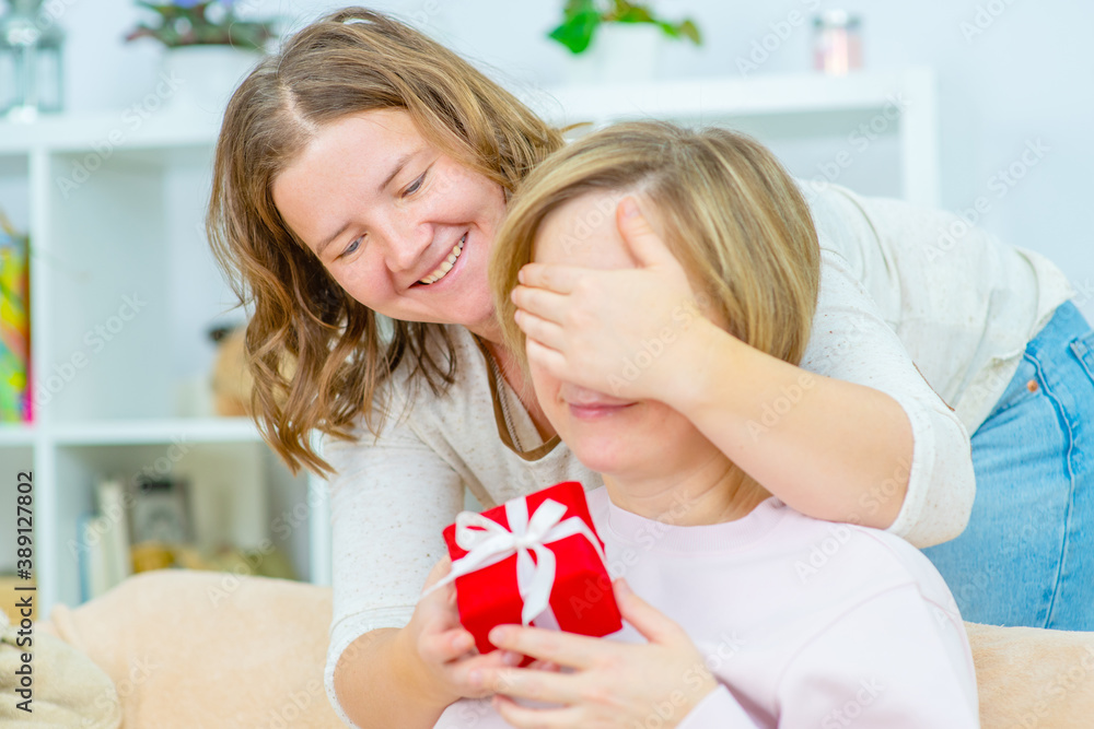 A young woman giving her friend a gift and hugging her, closing her eyes with her palm. Gay lesbian couple at home. Lesbian couple concept A young woman giving her friend a gift and hugging her, closing her eyes with her palm. Gay lesbian couple at home. Lesbian couple concept