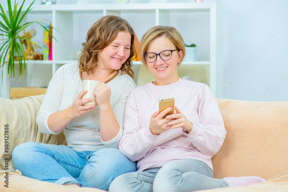 Two young women sit on the couch at home and look at the phone while having morning coffee. Gay lesbian couple at home. Lesbian couple concept Two young women sit on the couch at home and look at the phone while having morning coffee. Gay lesbian couple at home. Lesbian couple concept