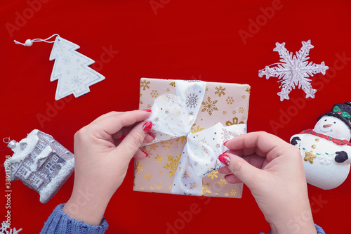 female hands are wrapping a gift, tying a ribbon on a red background with Christmas decorations.