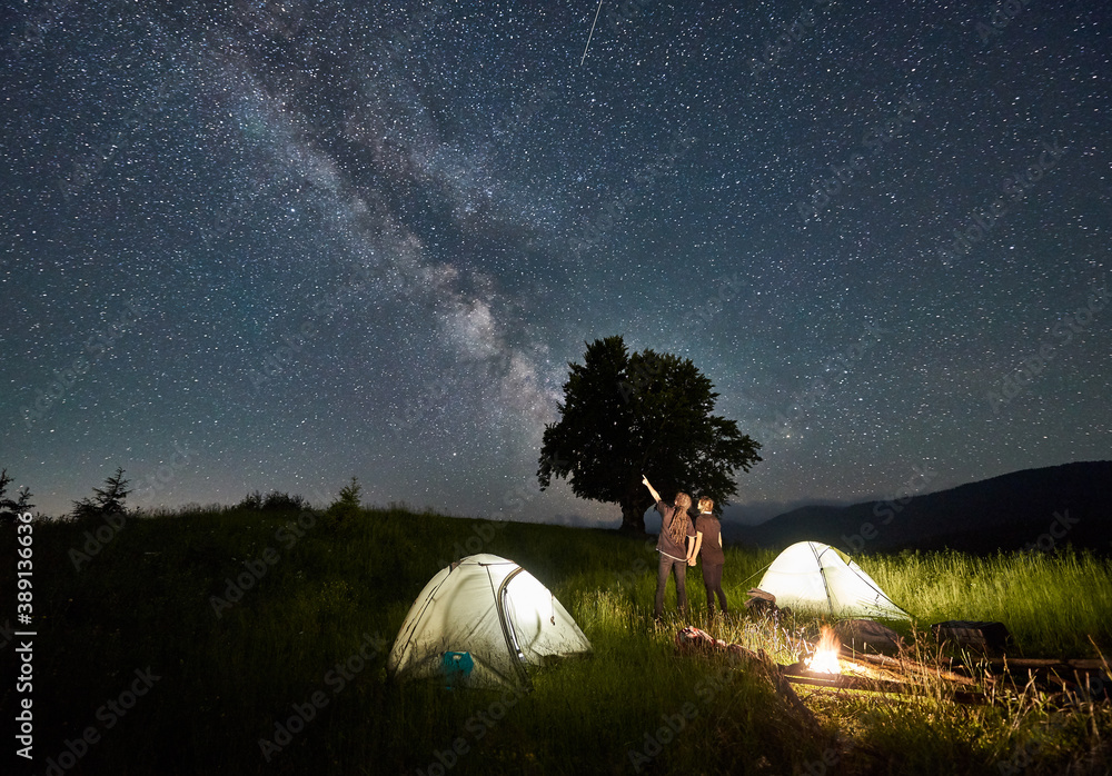 Man hiker with girlfriend pointing at Milky Way on majestic starry sky. Couple in love standing near illuminated camp tents, looking at sky with stars. Concept of hiking, night camping, relationships - obrazy, fototapety, plakaty 