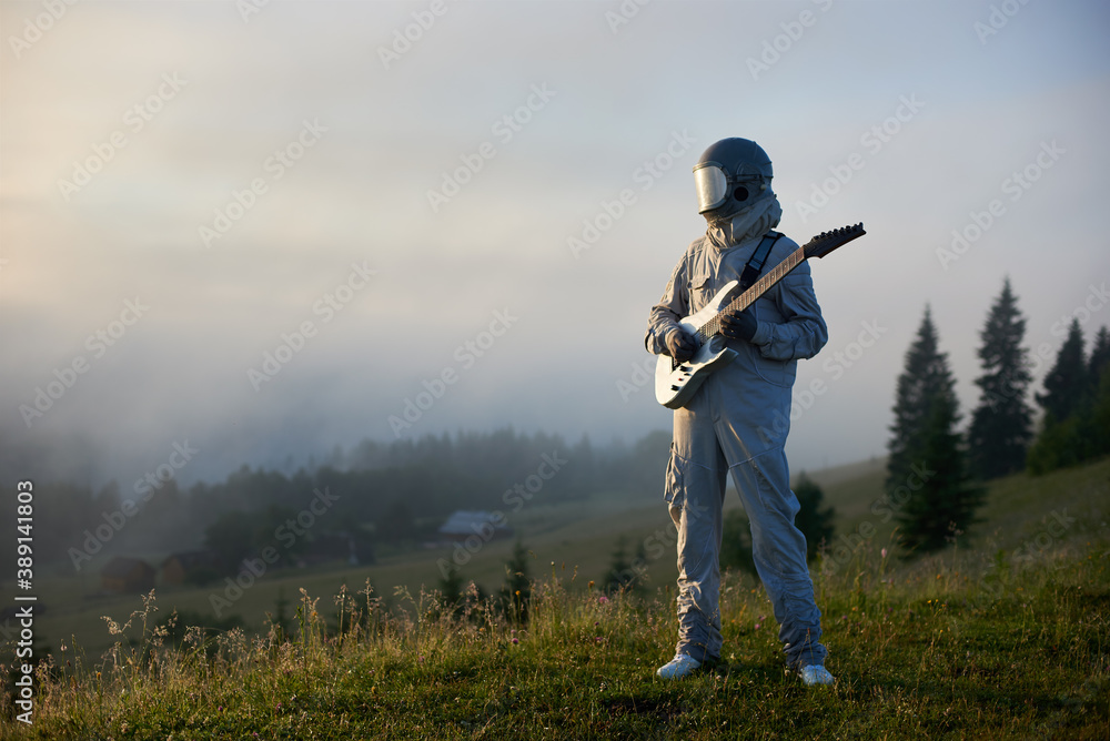 custom made wallpaper toronto digitalAstronaut guitarist in white space suit and helmet playing melody on guitar, standing in grassy meadow with foggy mountain village and sky on background. Concept of music, astronautics and nature.