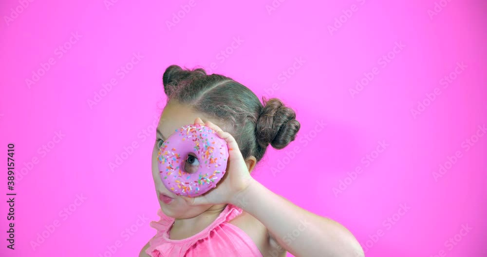Attractive cute little girl holding donuts with icing to her hands smiling looks at the camera and brings them to her face looking to look at collection of sweet donuts with pink icing. Portrait child Attractive cute little girl holding donuts with icing to her hands smiling looks at the camera and brings them to her face looking to look at collection of sweet donuts with pink icing. Portrait child