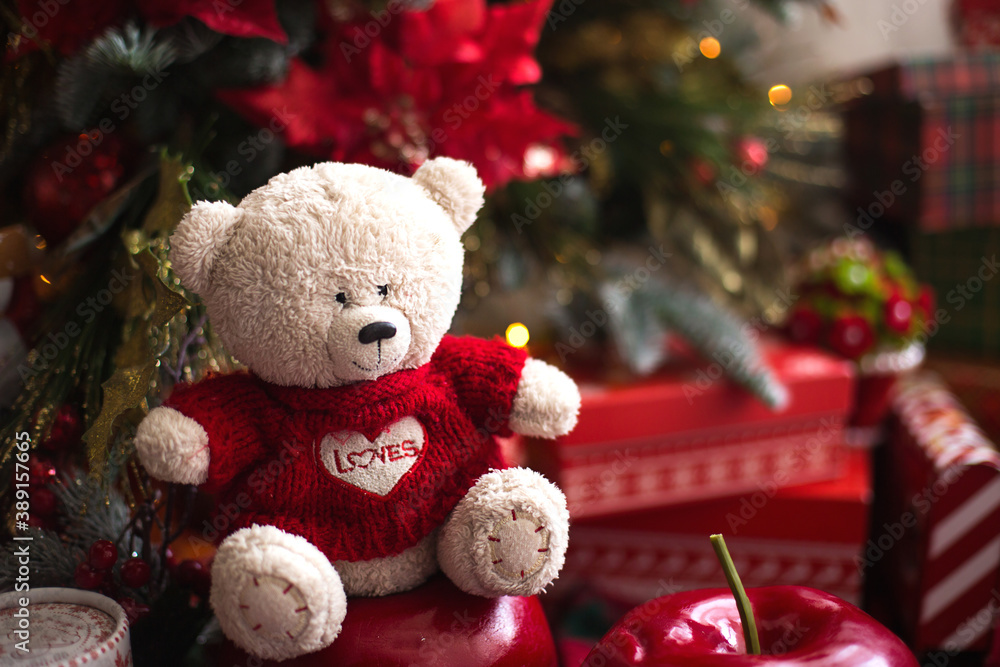 White Teddy bear in red knitted sweater with heart on the chest and the words Love near Christmas tree among the gift boxes. Gift to your beloved, Declaration of love, poinsettia decor. Copy space White Teddy bear in red knitted sweater with heart on the chest and the words Love near Christmas tree among the gift boxes. Gift to your beloved, Declaration of love, poinsettia decor. Copy space