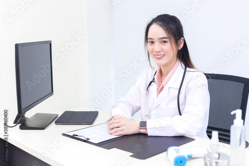Asian young woman doctor in a white lab coat is sitting on office room at hospital while has a computer, clipbroad and alcohol gel on the deck.