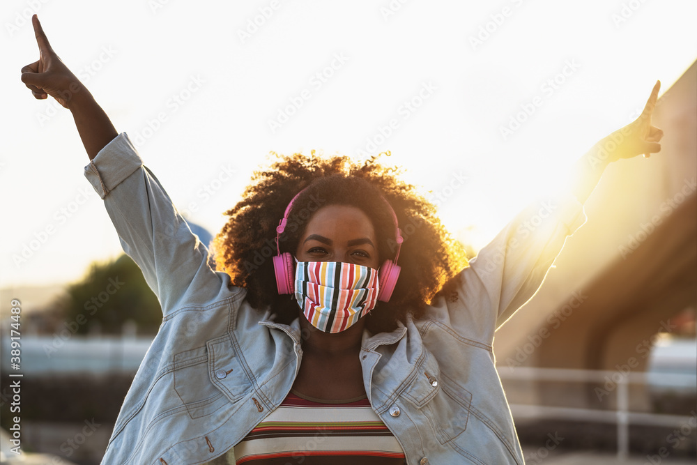 Happy Afro woman listening to playlist music with wireless headphones while wearing face colored mask outdoor - obrazy, fototapety, plakaty 