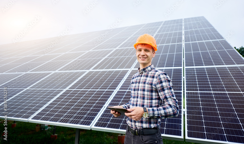 Smiling man in helmet with tablet in hands stands near solar panels on green plantation. Concept ecology protection. Man worker. Science technology. Smiling man in helmet with tablet in hands stands near solar panels on green plantation. Concept ecology protection. Man worker. Science technology.