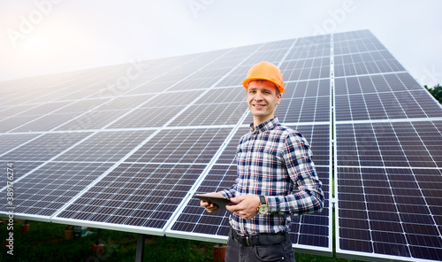 Smiling man in helmet with tablet in hands stands near solar panels on green plantation. Concept ecology protection. Man worker. Science technology.