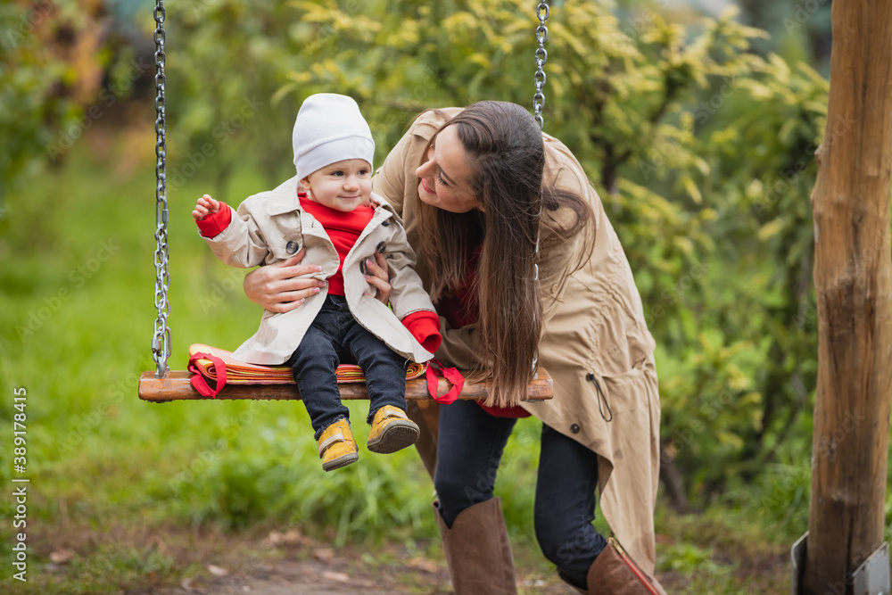 Fototapeta premium mother shakes her baby on swing at the autumn park. Happy family spend time together.