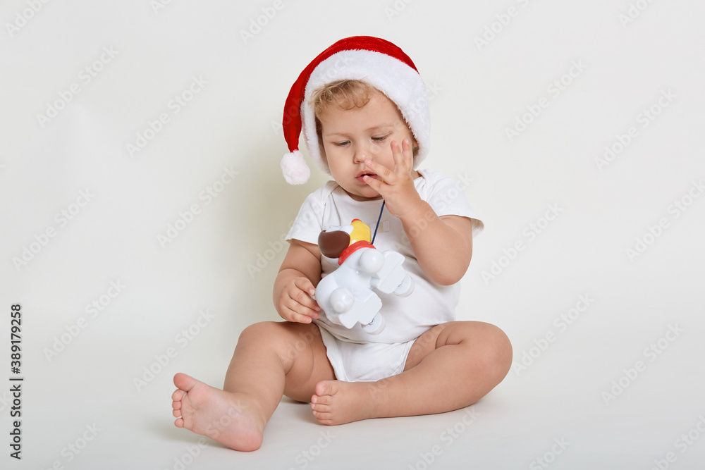 Sweet child, boy playing with plastic dog, sitting on floor against white background, being barefoot, wearing bodysuit and christmas hat, infant isolated over white wall. Sweet child, boy playing with plastic dog, sitting on floor against white background, being barefoot, wearing bodysuit and christmas hat, infant isolated over white wall.