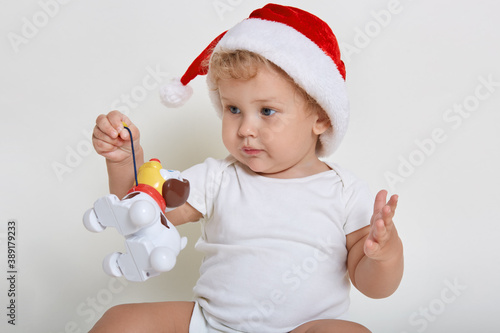 Cute baby wearing christmas hat and body suit playing with plastic dog, charming infant holding his toy, child looking away, baby looking away, sees new interesting toy aside.