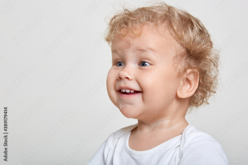 Little baby boy with curly blond hair laughing while looking attentively away wile posing isolated over white background, excited male child sees something interesting, copy space for promo.