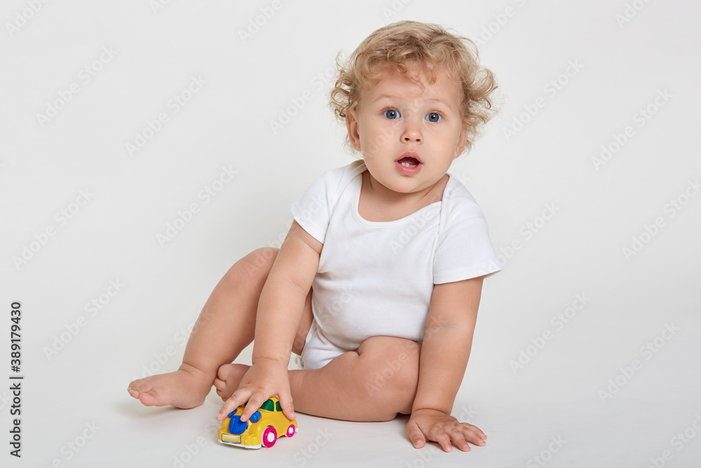 Portrait of adorable baby boy sitting on floor bare foot and dresses body suit, looking in camera with opened mouth and showing his teeth, kid playing colored toy car. Portrait of adorable baby boy sitting on floor bare foot and dresses body suit, looking in camera with opened mouth and showing his teeth, kid playing colored toy car.