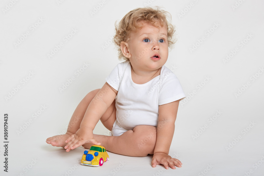 Funny baby boy playing toy in nursery or kindergarten, curly haired blond infant in body suit sitting on floor bare foot, child looking away with curious look, posing isolated over white background. Funny baby boy playing toy in nursery or kindergarten, curly haired blond infant in body suit sitting on floor bare foot, child looking away with curious look, posing isolated over white background.