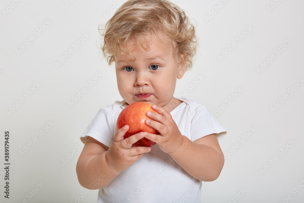 Baby boy holding and eating red apple, posing isolated on white background, charming male child with blond curly hair looking away, looks concentrated, wearing t shirt. Baby boy holding and eating red apple, posing isolated on white background, charming male child with blond curly hair looking away, looks concentrated, wearing t shirt.