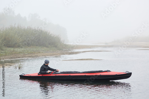 Side view of male kayaking on river, sportsman in black jacket looking at foggy water, holds oar in hands, foggy day, man in boat with bank of river and bushes on background.