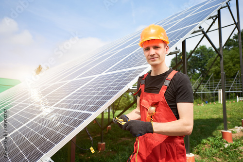 Male worker in orange uniform and black gloves with equipment in hands working at solar station, smiling to the camera. On background of solar panels. Home construction