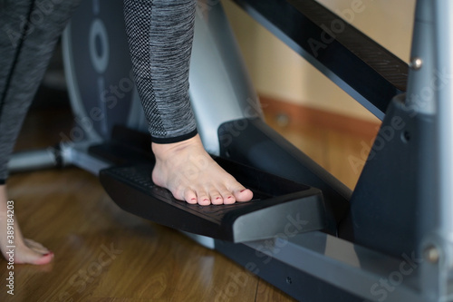 A barefoot girl stands on the pedal of an ellipsoid and begins training. Close-up of legs without face. Fitness machine and sports at home photo