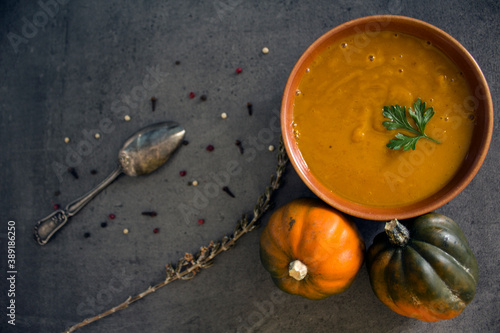 Top view photo of bowl with pumpkin soup, gem squashes, rosemary, pepper and silver spoon. Dark grey textured background. Healthy eating concept. Autumn menu. 