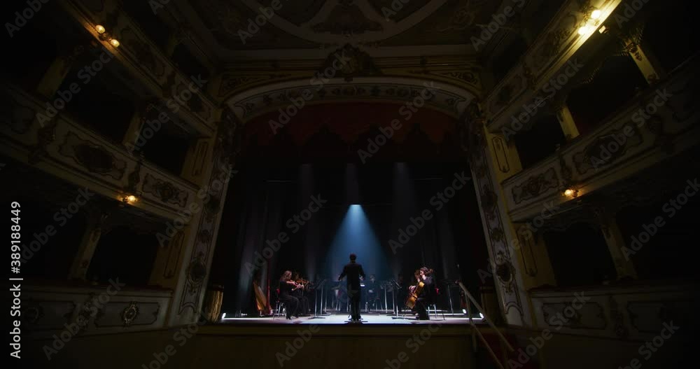 Cinematic shot of conductor directing symphony orchestra with performers playing violins, cello and trumpet on classic theatre with curtain stage during music concert with dramatic lights. Cinematic shot of conductor directing symphony orchestra with performers playing violins, cello and trumpet on classic theatre with curtain stage during music concert with dramatic lights.