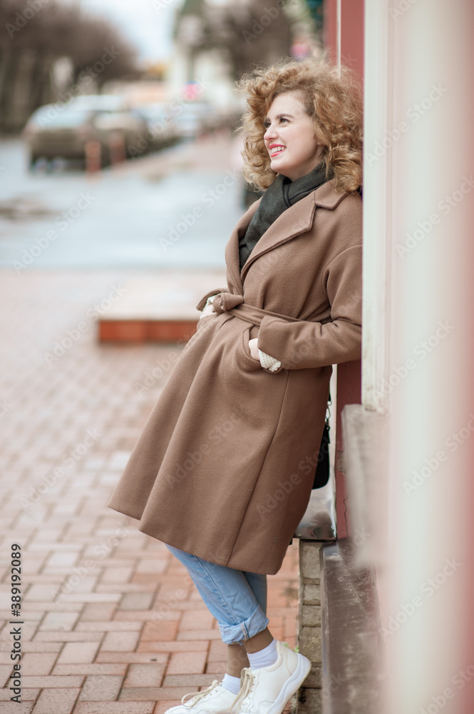 Portrait of a smiling young woman with curly hair. Outside. Girl standing leaning against the wall