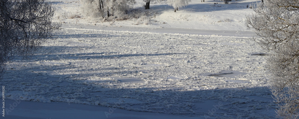 White icy snow covered frosty river surface after snowfall with trees on riverbank, beautiful European countryside landscape at Sunny winter day White icy snow covered frosty river surface after snowfall with trees on riverbank, beautiful European countryside landscape at Sunny winter day