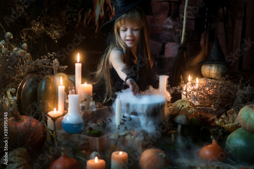 A cute little witch cooks a potion over a bonfire of herbs and mushrooms. Beautiful baby girl in witch costume with Halloween decoration