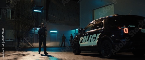 Фотография WIDE Police officer exiting a car and approaching two multi-ethnic males suspects near industrial buildings at night
