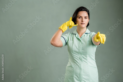Young cleaning woman wearing a green shirt and yellow gloves showing call me gesture, pointing her finger © Cipri Suciu 
