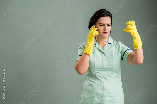Young cleaning woman wearing a green shirt and yellow gloves displeased face and gestures and talking on cellphone © Cipri Suciu 