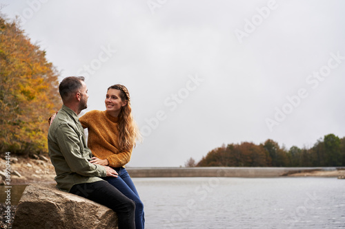 Couple sitting in front of the lake and looking at each other outdoors with a lake in the background.