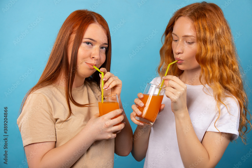 Red-haired girlfriends in casual clothes are drinking orange juice with straws. Isolated on a blue background. People lifestyle concept. Red-haired girlfriends in casual clothes are drinking orange juice with straws. Isolated on a blue background. People lifestyle concept.