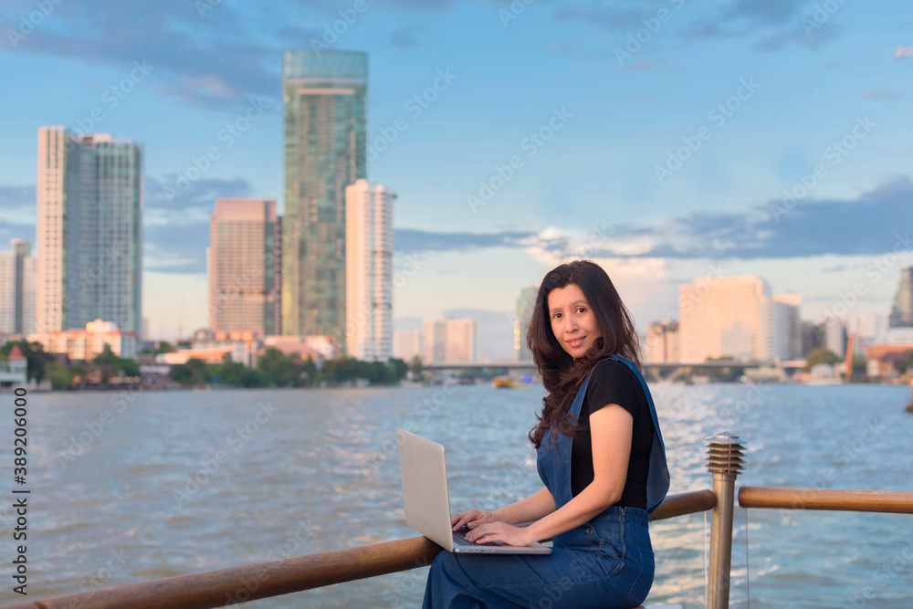 Asian woman using laptop in front of river beautiful view at sunset and background blur building skyscrapers. (Technologies and business concept, copy space)