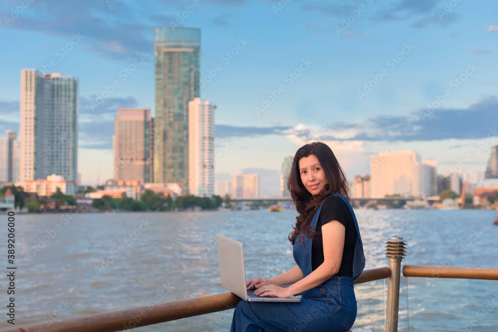 Asian woman using laptop in front of river beautiful view at sunset and background blur building skyscrapers. (Technologies and business concept, copy space) Asian woman using laptop in front of river beautiful view at sunset and background blur building skyscrapers. (Technologies and business concept, copy space)