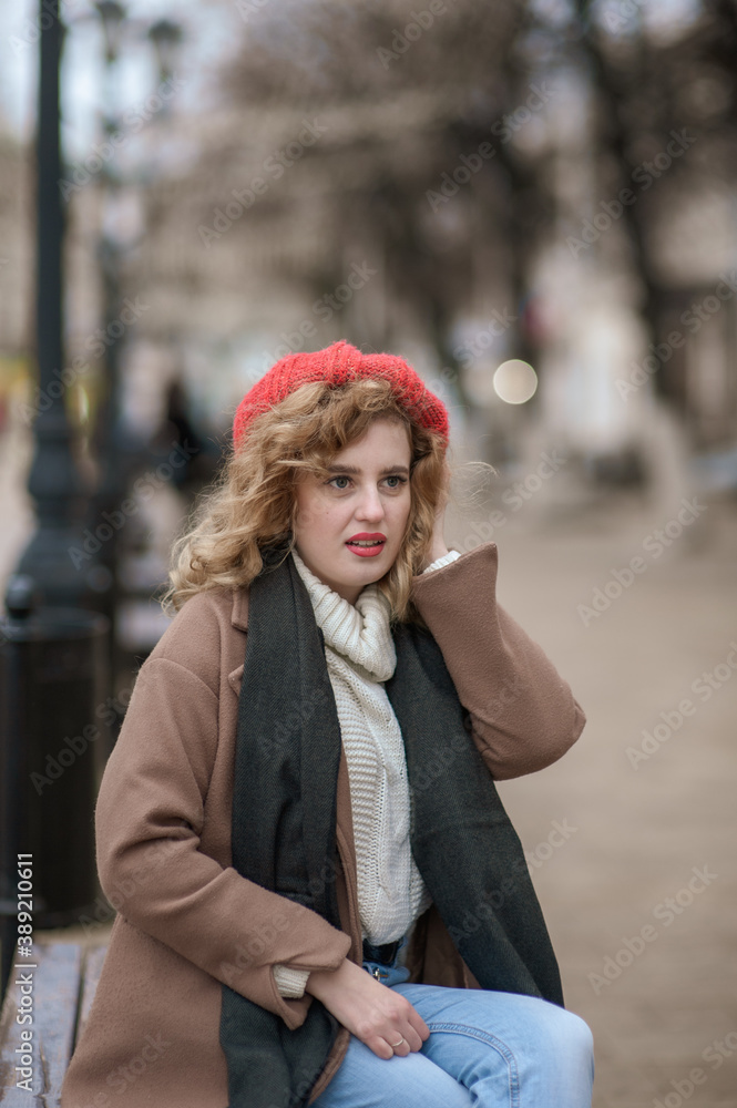 A cute girl in a red hat sits on a wooden bench on a city street A cute girl in a red hat sits on a wooden bench on a city street