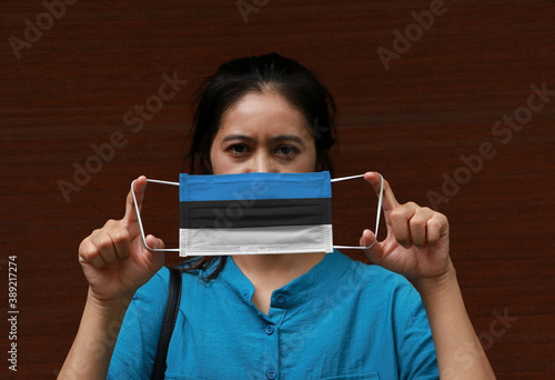 A woman and hygienic mask with Estonia flag pattern in her hand and raises it to cover her face on green background.