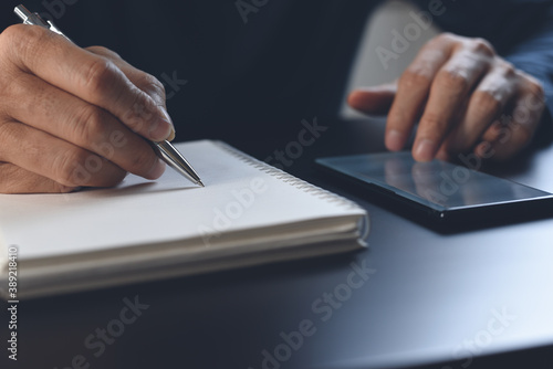 Close up of man hand writing on paper notebook and using mobile phone on table