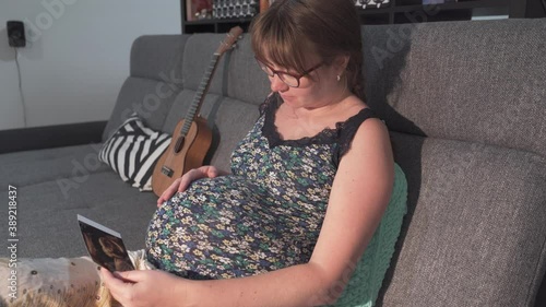 A pregnant woman is sitting on the sofa at home, looking at the ultrasound picture of her unborn baby and caressing her belly. Ninth month of pregnancy.