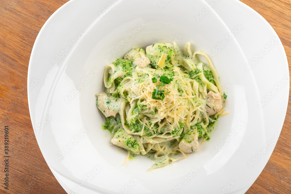 Tagliatelle pasta with pesto, chicken and Parmesan in a white plate on a wooden table, top view