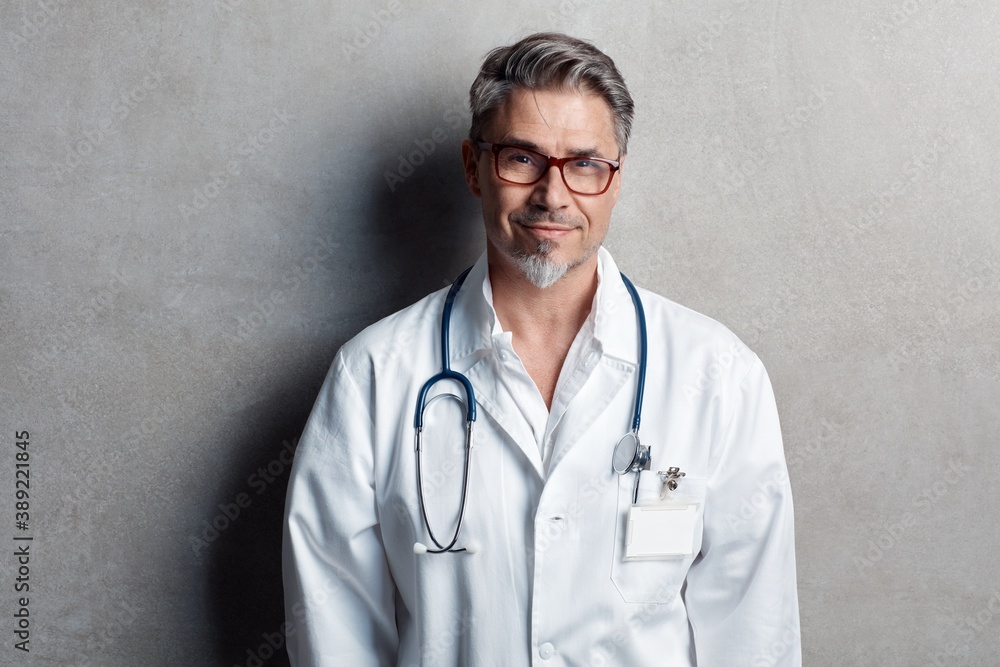 Portrait of trustworthy older smart doctor with gray hair wearing glasses and white lab coat standing against gray wall, smiling. Copy space. Portrait of trustworthy older smart doctor with gray hair wearing glasses and white lab coat standing against gray wall, smiling. Copy space.
