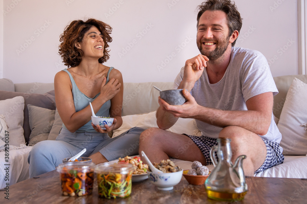 Couple eating healthy food from a delivery on the couch smiling Couple eating healthy food from a delivery on the couch smiling