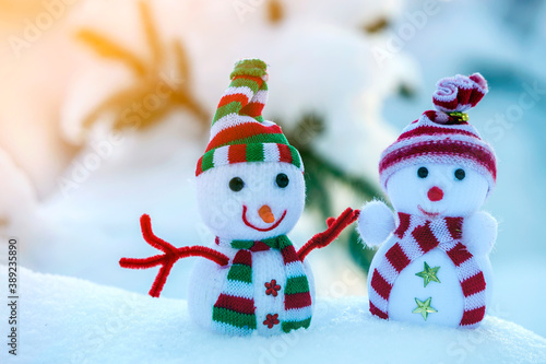 Two small funny toys baby snowman in knitted hats and scarves in deep snow outdoors near pine tree branch. Happy New Year and Merry Christmas greeting card.